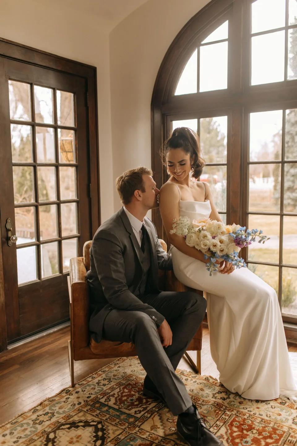 Bride holding a bouquet sits beside her partner in a sunlit room